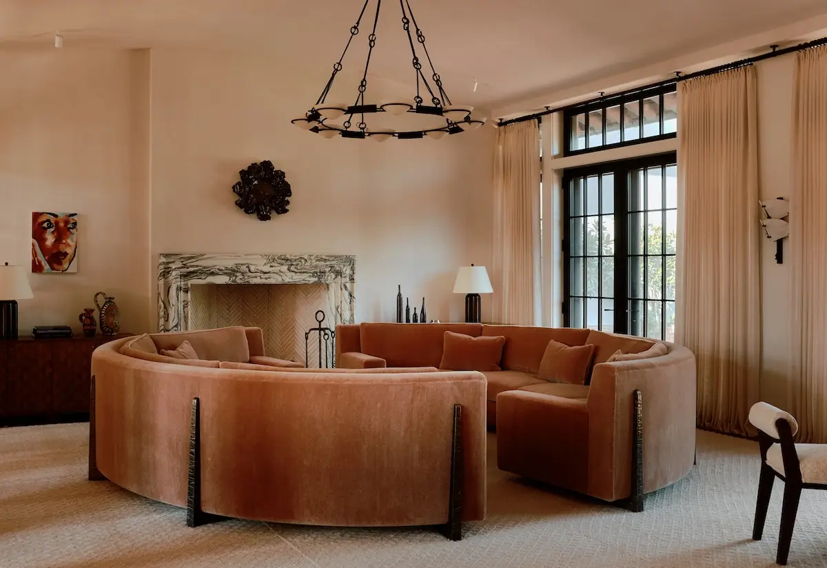 Contemporary living room with a curved rust-colored velvet sectional sofa, cream walls, marble fireplace, black-framed windows, and warm neutral decor.