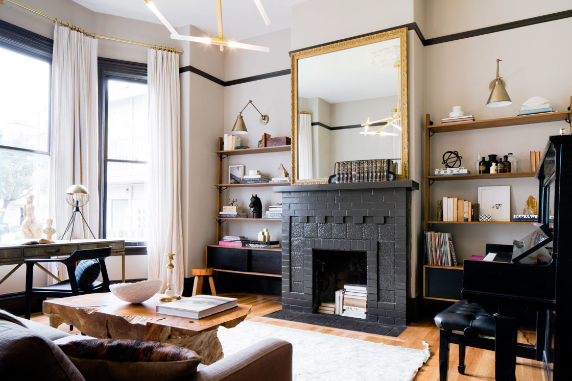 Modern Victorian living room with a black-painted original fireplace, gold-framed mirror, custom open shelving, tall windows with white curtains, and contemporary furniture.