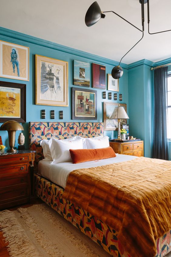Eclectic bedroom features bold turquoise blue walls where framed art creates a gallery wall above the bed frame, which is covered in a vibrant print 