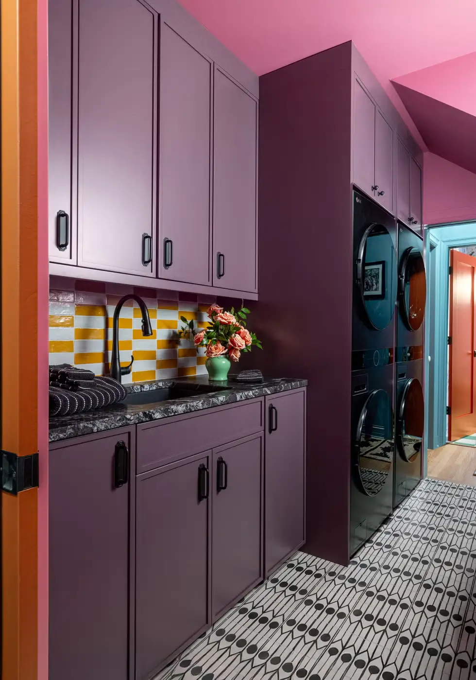 Laundry room painted in mauve and pink with patterned black-and-white floor tiles, striped yellow backsplash, and black appliances.