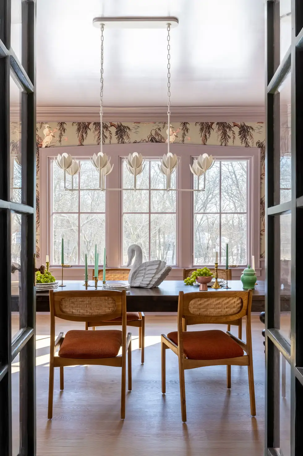 Dining room with blush pink walls, large bay windows, mid-century wood chairs, floral wallpaper, and white lotus pendant chandelier.