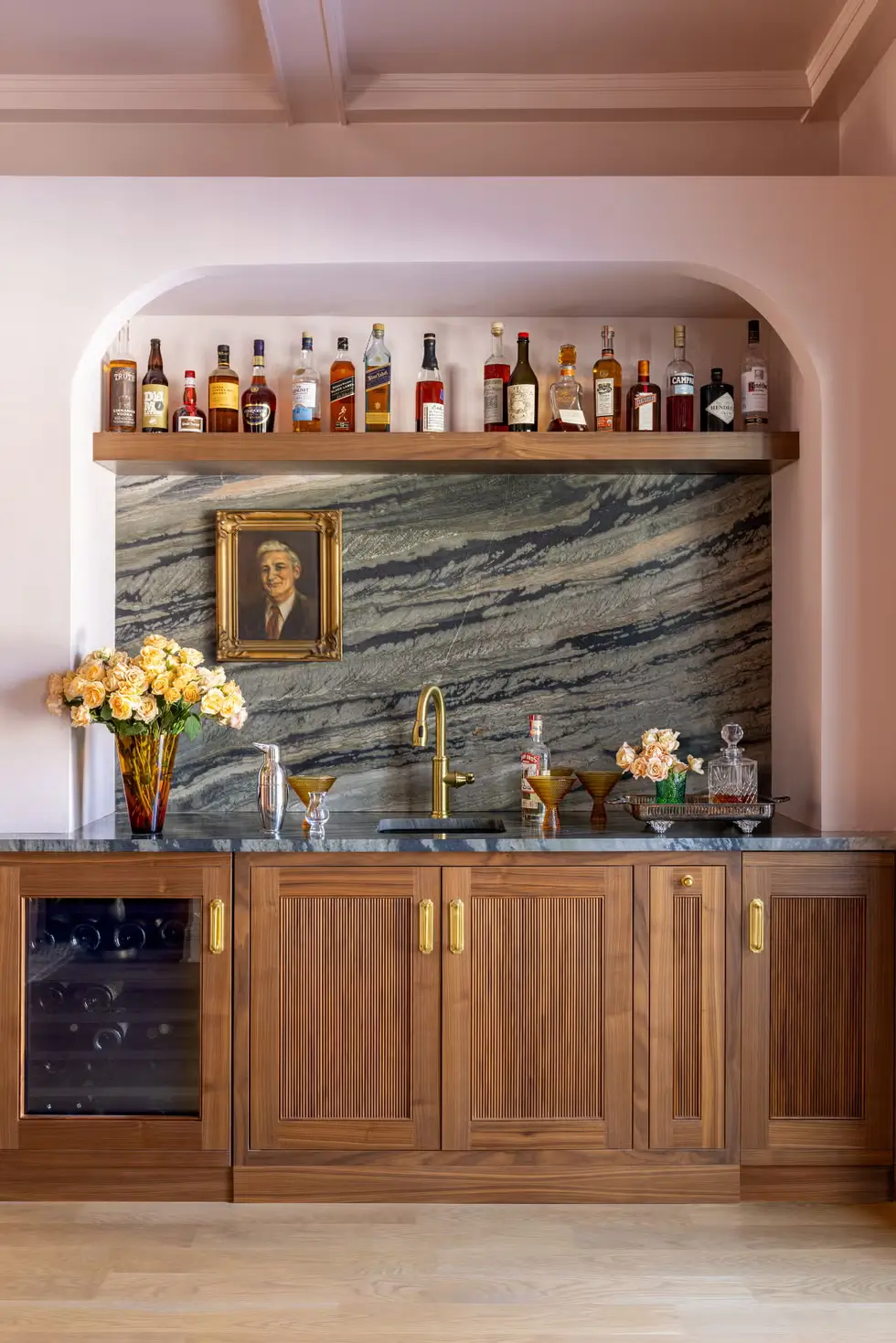 Home bar area with walnut cabinetry, marble backsplash, brass fixtures, and a portrait above the sink in a playful vintage-meets-modern farmhouse.