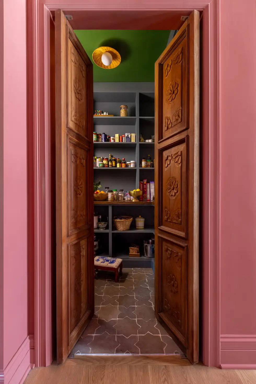 Pantry with carved antique wooden doors, pink hallway walls, emerald green ceiling, and gray shelving filled with colorful pantry items.