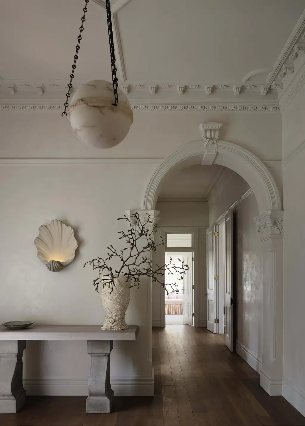A serene, minimalist hallway interior with classical architectural detailing and a soft neutral palette. The space features ornate crown molding, decorative archways, and warm wooden floors that contrast elegantly with creamy white walls. A sculptural marble pendant light hangs from black chains, while a shell-shaped wall sconce adds a subtle artistic touch. A stone console table styled with handcrafted ceramics and twisting branches creates an organic focal point. The overall atmosphere feels timeless, refined, and quietly luxurious, blending European heritage architecture with contemporary minimal styling and natural textures.
