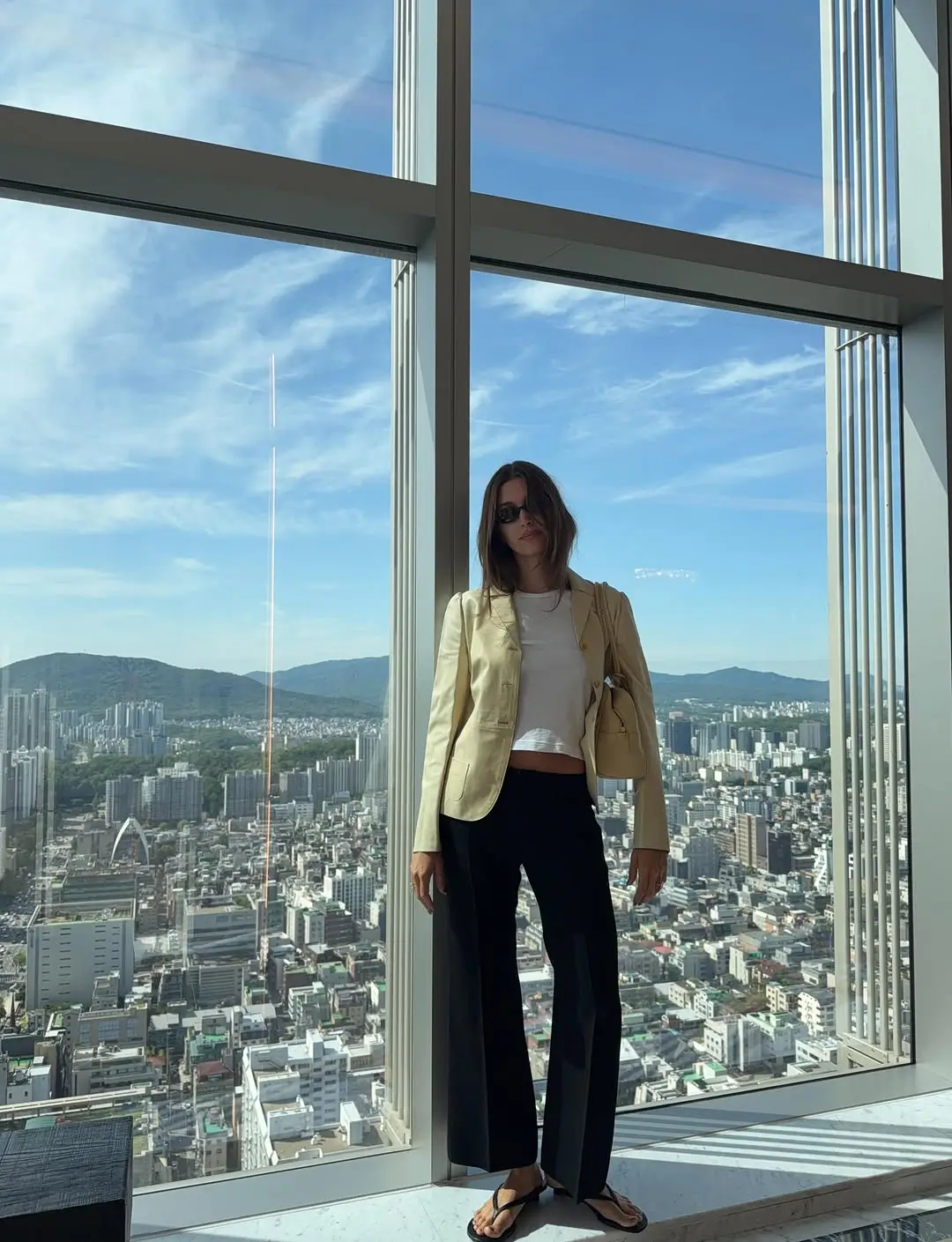 Hailey Bieber wears black minimalist flip flops with tailored black pants, a white t-shirt, and a cream jacket while posing in front of the city skyline in Seoul.