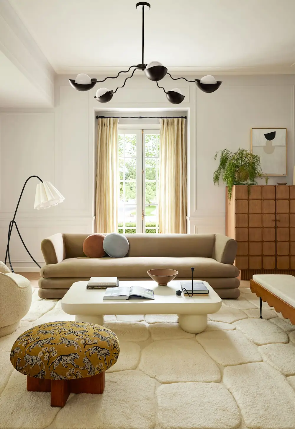 A bright, refined living room blending contemporary softness with subtle mid-century influences. The space features a sculptural beige sofa with rounded layered upholstery, a plush geometric cream rug, and a minimalist ivory coffee table with chunky cylindrical legs. Black sculptural lighting fixtures contrast elegantly against the soft neutral palette, while warm wood accents and textured fabrics add depth and warmth. Large French windows with soft drapery flood the room with natural light, creating an airy, serene atmosphere. Carefully curated decor, organic forms, and muted earthy tones give the interior a sophisticated yet inviting modern aesthetic.
