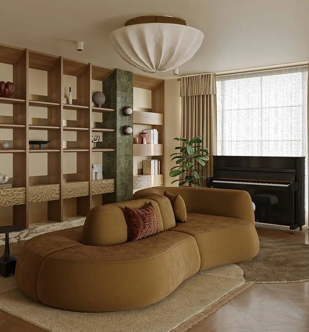 A warm contemporary living room with a sculptural curved caramel-brown sofa as the centerpiece, paired with rich wood shelving and soft neutral tones. The custom built-in shelving wall combines open oak compartments, marble surfaces, and a vertical strip of glossy green tiles for an architectural focal point. A large pleated fabric ceiling light adds softness overhead, while layered rugs, textured cushions, and floor-length drapery create a cozy atmosphere. The space blends organic modern design with mid-century influences, emphasizing rounded forms, earthy colors, natural materials, and refined minimalist styling.
