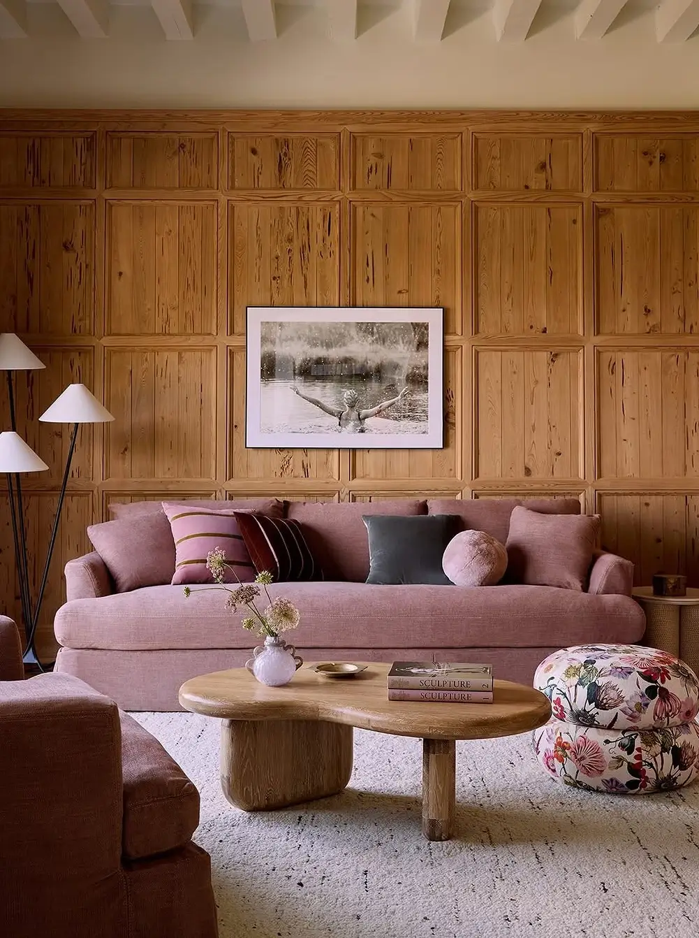 A cozy, softly feminine living room with warm wood-paneled walls and blush-toned furnishings creating an inviting, layered atmosphere. The space features a plush pink sofa styled with velvet and striped accent pillows, paired with a sculptural organic wood coffee table and a floral upholstered pouf. Natural wood textures, muted rose hues, and soft neutral textiles balance rustic warmth with contemporary comfort. Minimal black floor lamps and framed monochrome artwork add contrast, while the overall design blends Scandinavian-inspired simplicity with romantic vintage touches and earthy modern styling.