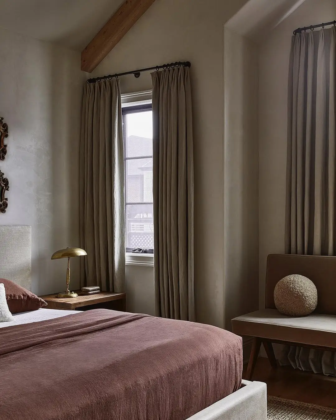 Warm earthy bedroom with taupe walls, soft beige curtains, natural wood accents, and muted mauve bedding. Layered neutral tones, textured fabrics, and warm lighting create a cozy, calming atmosphere with an understated luxury feel.