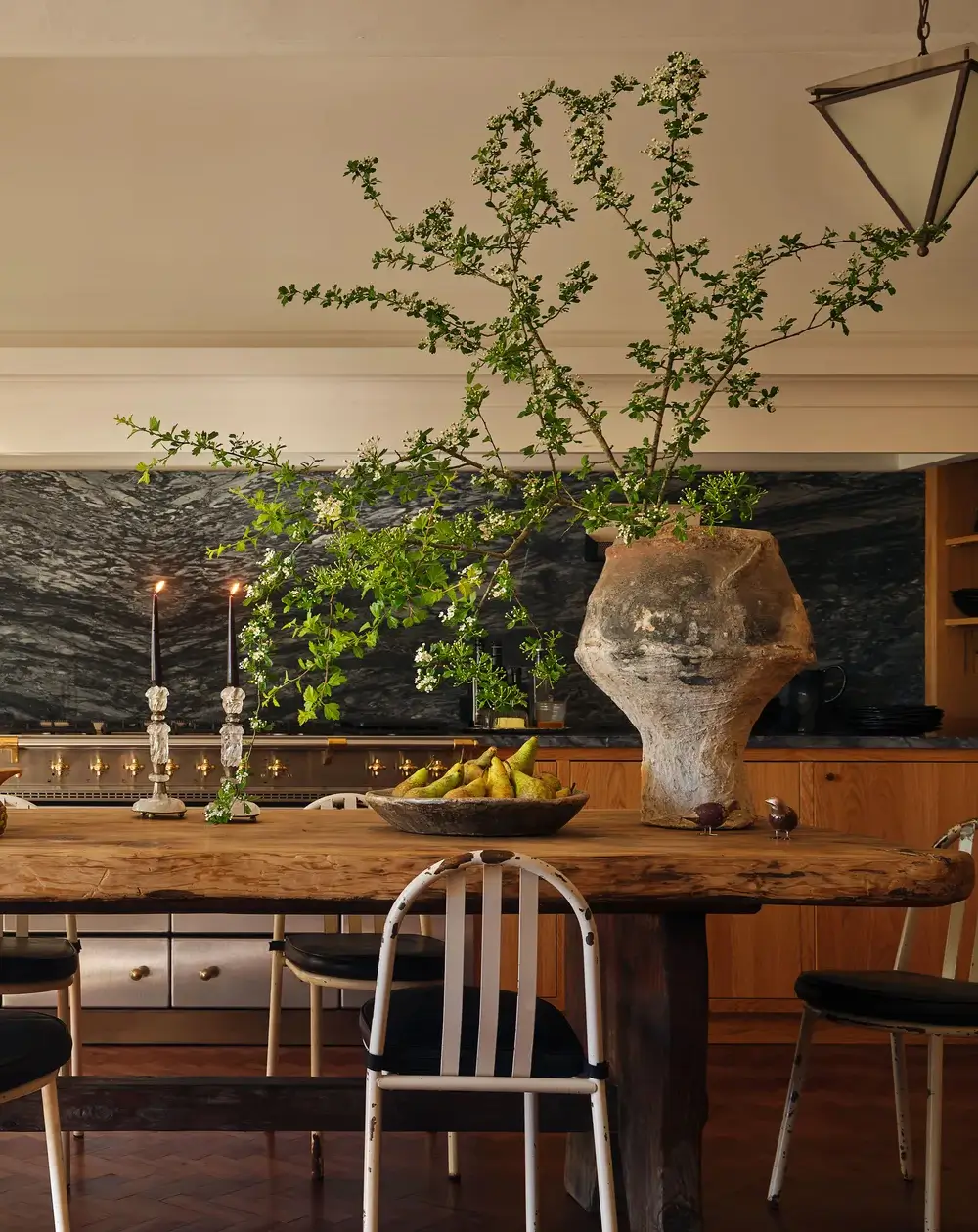 Collected cinematic kitchen with a raw-edge antique wood table, vintage painted metal chairs, dramatic dark marble backsplash, warm wood cabinetry, candlelight, and a large weathered clay vase filled with orchard branches creating a layered European-inspired atmosphere.