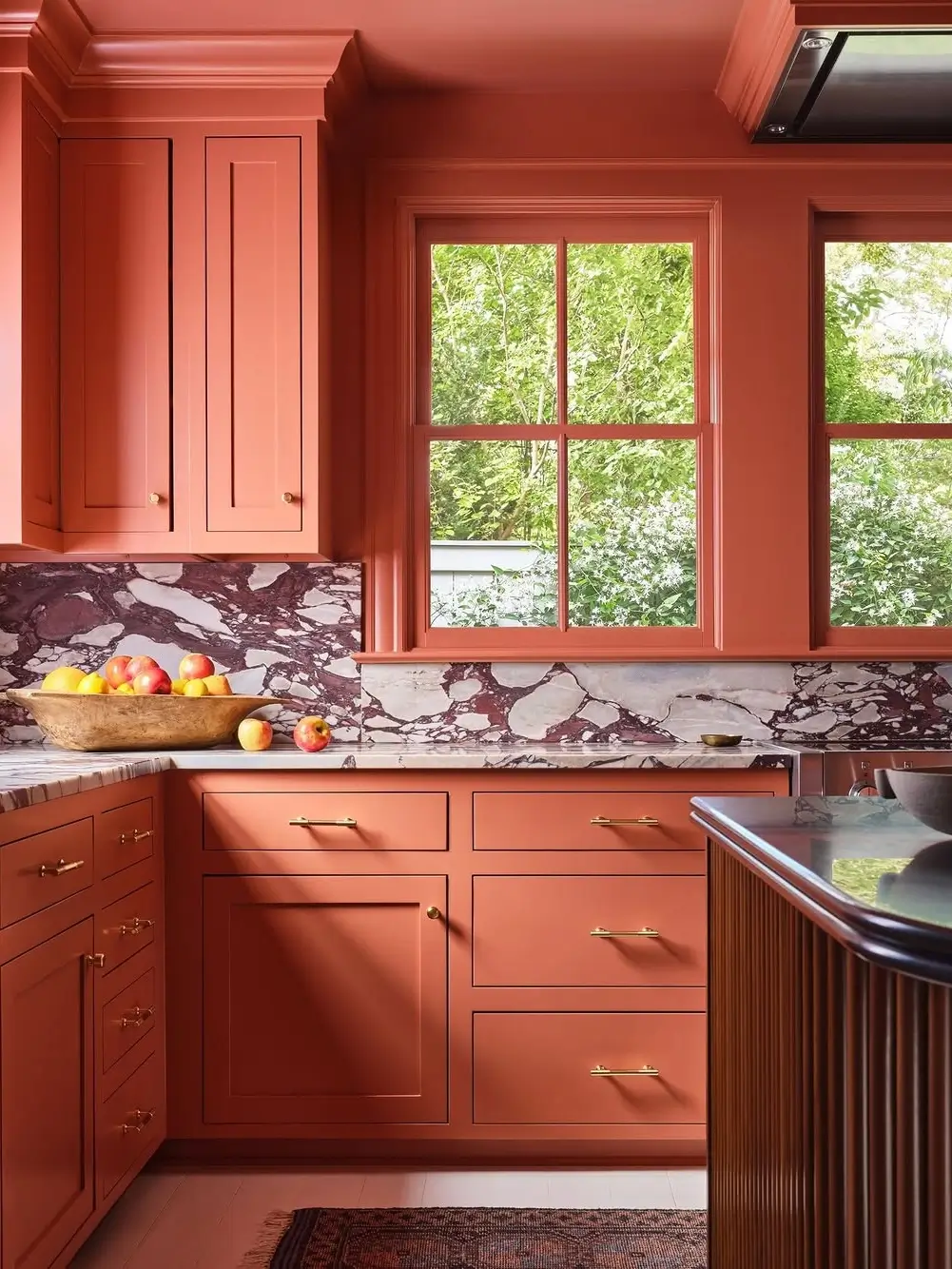 Terracotta cinematic kitchen with dramatic marble backsplash, tonal clay-colored cabinetry, brass hardware, fluted wood island detailing, and warm natural light creating a rich, atmospheric European-inspired interior.