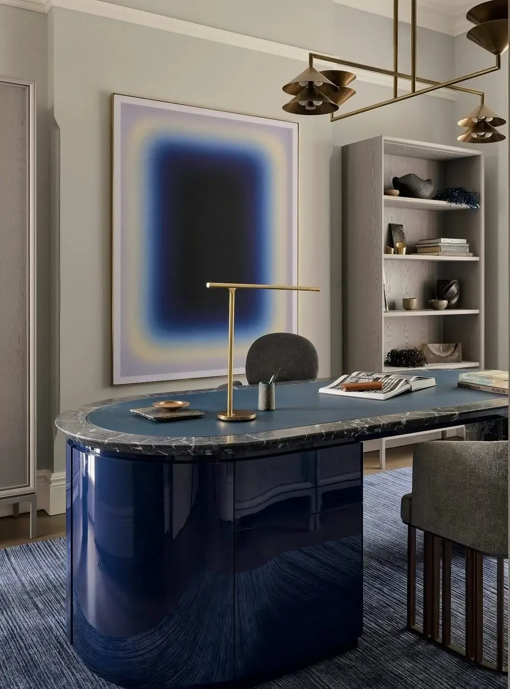 A sophisticated contemporary home office with a dramatic deep-blue lacquered desk featuring rounded edges and a dark marble top. The room combines muted greige walls, warm brass lighting, and minimalist styling for a refined modern aesthetic. A large abstract artwork with blurred blue gradients creates a bold focal point behind the desk, while open shelving displays curated ceramics, books, and sculptural decor. The layered textures of velvet, wood, marble, and brushed metal add depth and luxury, blending modern elegance with art-gallery-inspired minimalism.