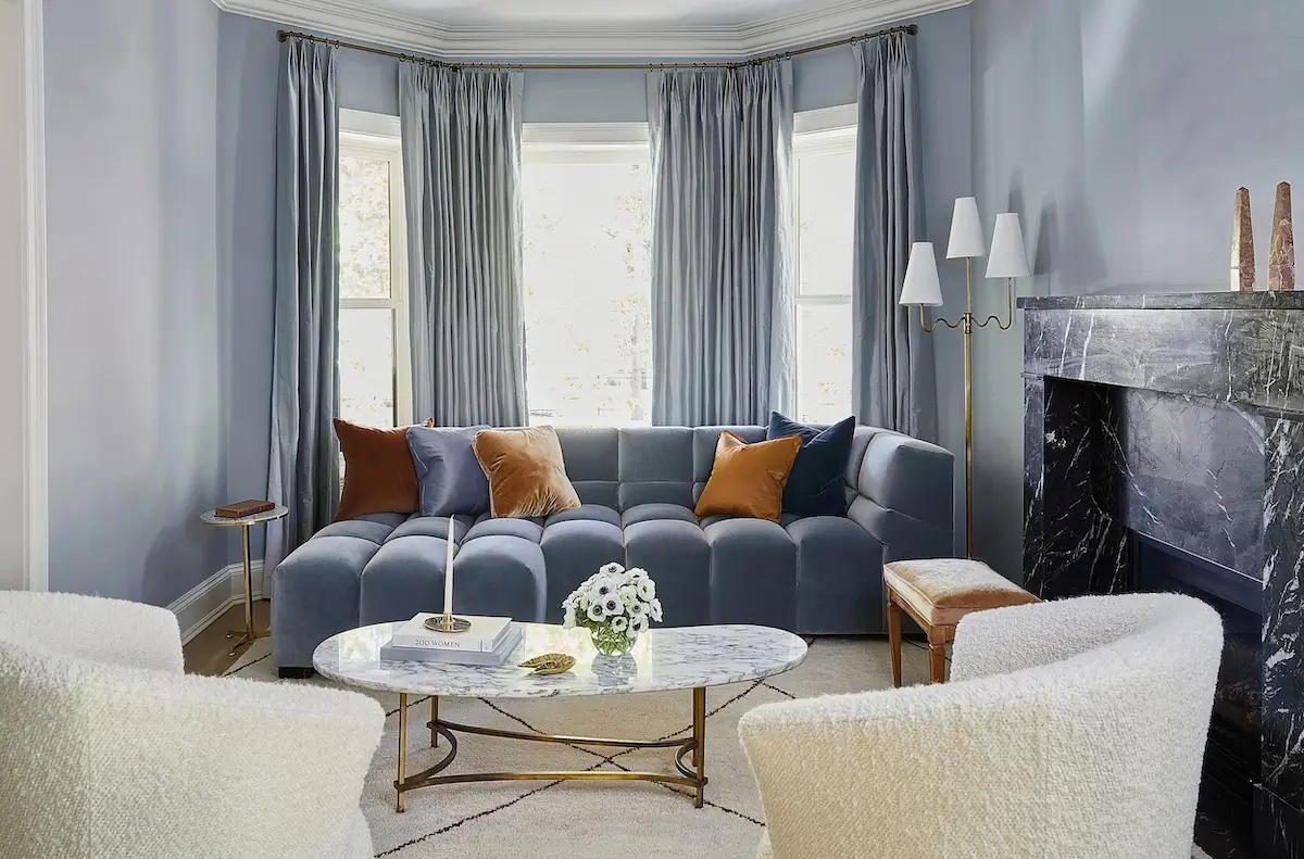 Soft blue-gray living room with a curved tufted velvet sofa, pale gray walls and curtains, boucle armchairs, and a black marble fireplace. Warm rust and navy accent pillows add contrast to the cool-toned palette, while brass details and layered textures create an elegant, modern atmosphere.