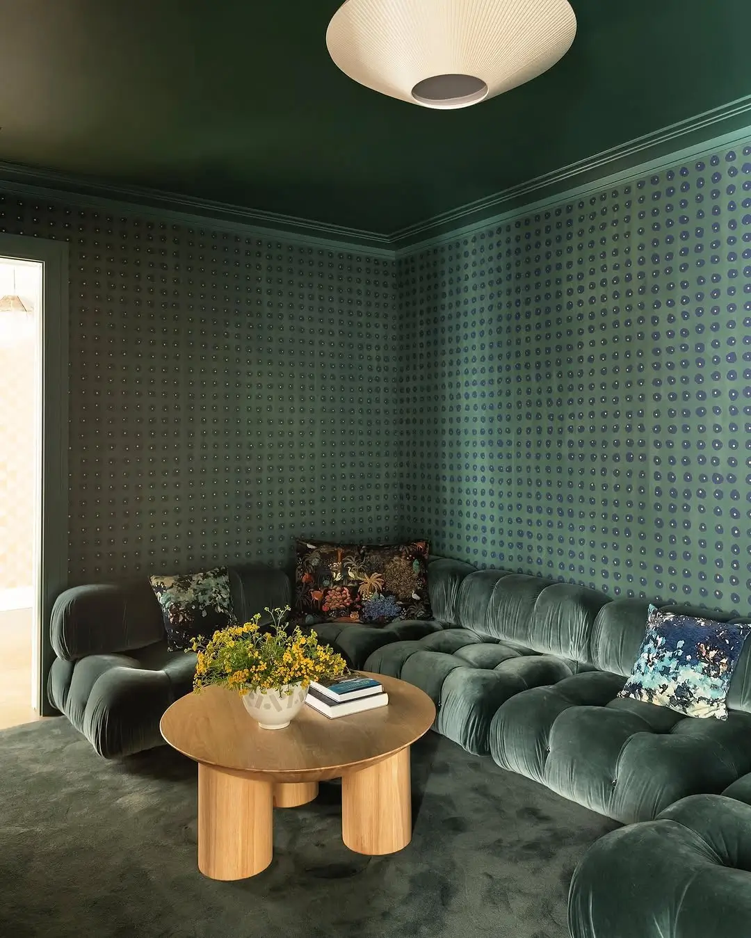 Moody green screening room designed by Studio DB featuring deep green hand-painted wallpaper, a matching tufted velvet Bellini-style sectional sofa, dark green carpet, floral accent pillows, and a round oak coffee table with yellow flowers. The cozy, color-drenched space has a warm, cinematic atmosphere with soft lighting and layered textures.