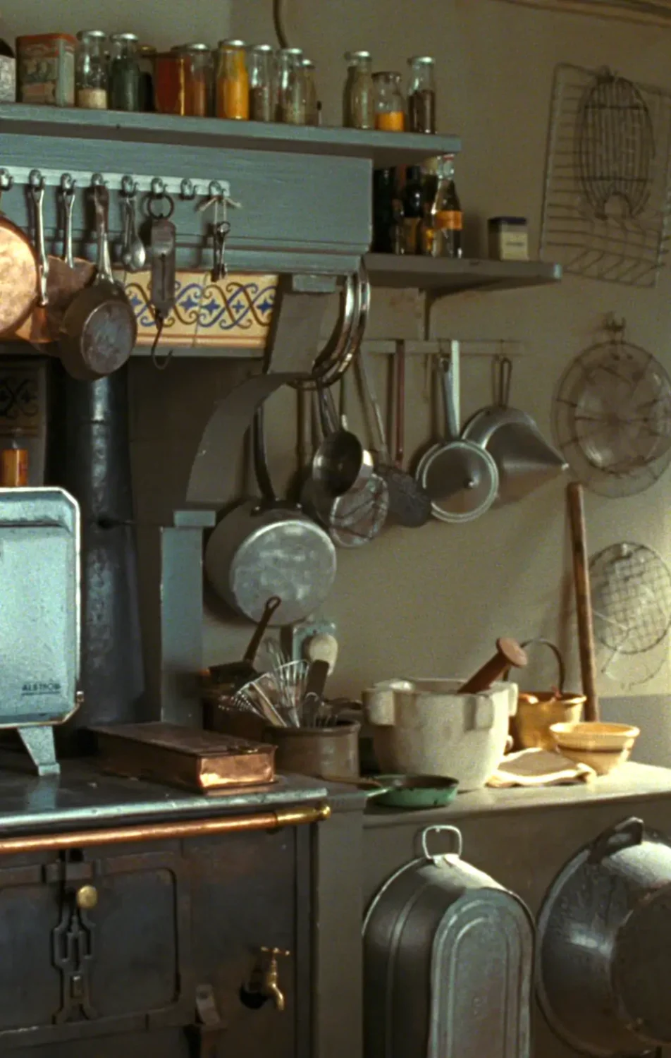 Close-up of the atmospheric kitchen from Julie & Julia, featuring hanging copper and metal cookware, open shelves lined with spice jars, vintage French cooking utensils, moody lighting, and an old cast-iron stove with beautifully lived-in European kitchen details.