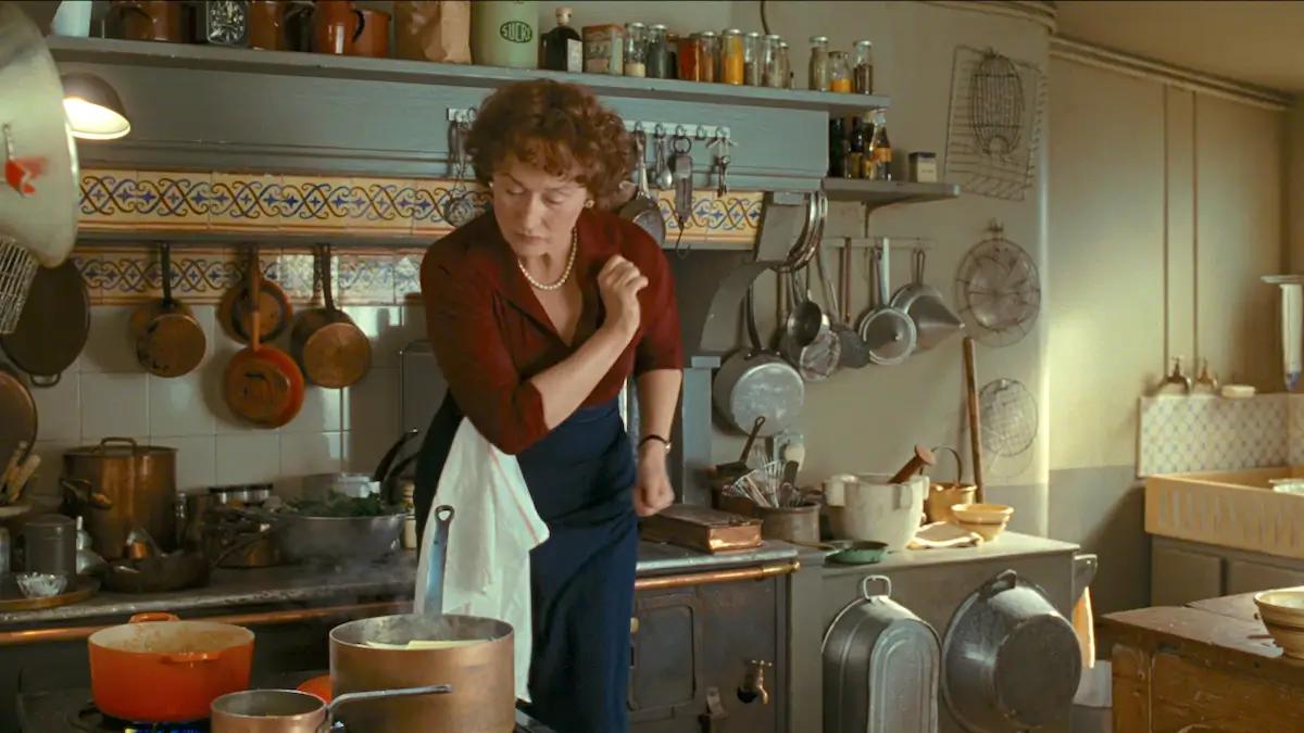 Meryl Streep as Julia Child cooking in the warm, atmospheric Paris kitchen from Julie & Julia, featuring hanging copper cookware, open shelving, vintage utensils, moody lighting, and layered old-world French kitchen details.