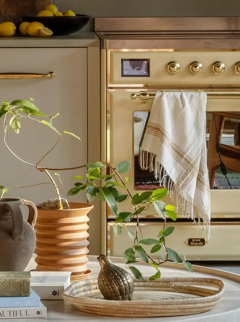 Close-up of a butter-yellow ILVE Majestic range cooker with brass knobs and handles, layered textiles, warm neutral cabinetry, ceramic decor, natural greenery, and soft sunlight creating a cozy cinematic kitchen atmosphere.