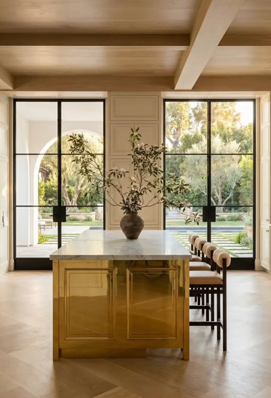 Warm luxury kitchen with brass island cabinetry, marble countertop, upholstered bar stools, and indoor-outdoor connection to garden