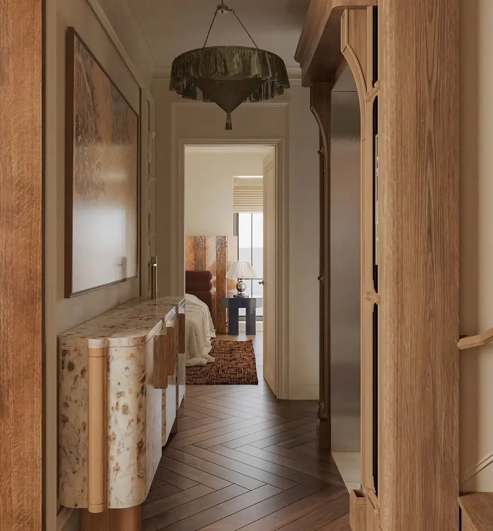 A warm, elegant hallway with rich wood textures, herringbone flooring, and softly layered neutral tones. The space features sculptural custom cabinetry, a curved floral stone console table, and a dramatic fringed pendant light that adds vintage-inspired character. Warm ambient lighting and natural materials create an intimate, luxurious atmosphere, while the view into the softly styled bedroom enhances the sense of depth and calm. The design blends contemporary craftsmanship with Art Deco and European-inspired details, emphasizing refined textures, organic forms, and understated sophistication.