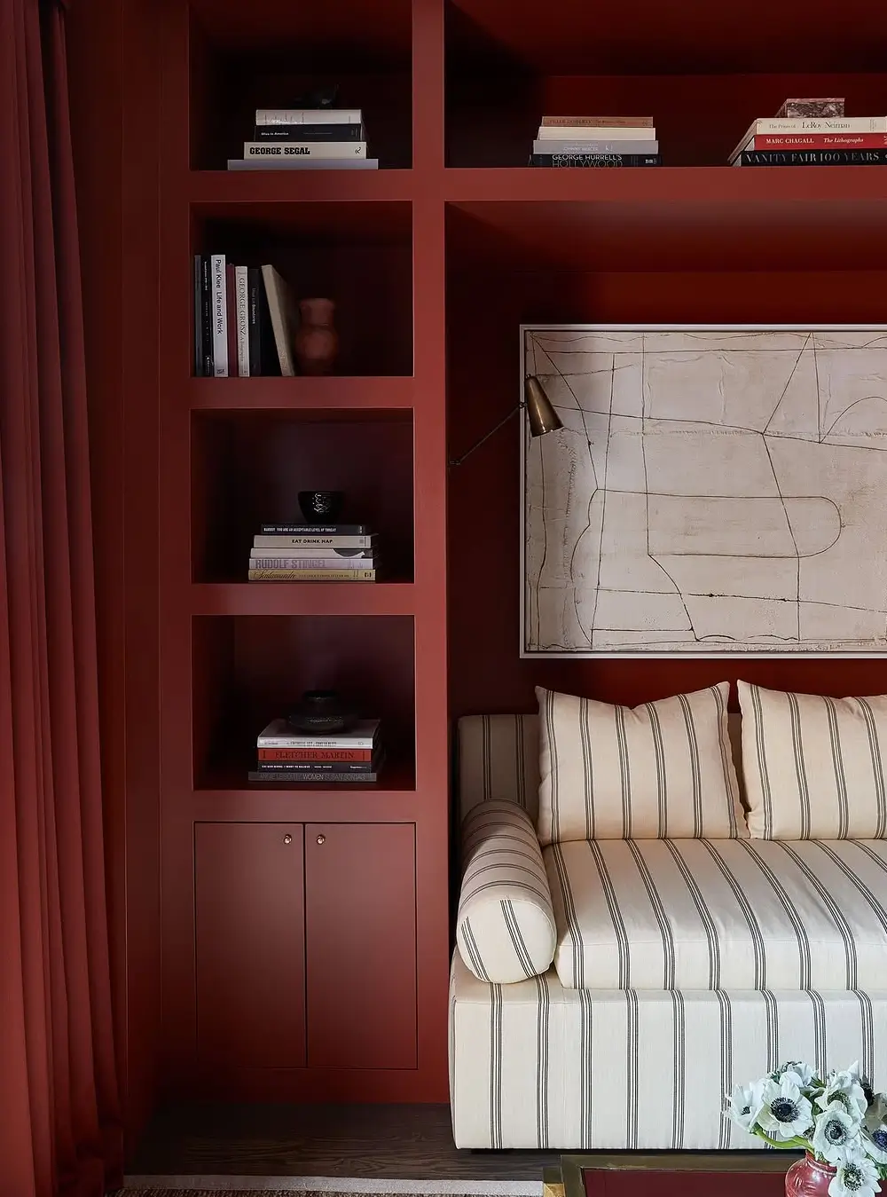 A dramatic, moody interior with deep oxblood-red built-in shelving and walls creating a rich monochromatic backdrop. The space features custom floor-to-ceiling cabinetry with open display niches styled with art books, ceramics, and minimal decor. A cream striped upholstered sofa softens the dark palette, paired with an abstract neutral-toned artwork and a brass wall-mounted reading lamp. Heavy burgundy curtains and dark wood flooring enhance the intimate, luxurious atmosphere. The design blends contemporary sophistication with vintage-inspired warmth, emphasizing bold color blocking, tailored millwork, and curated styling.