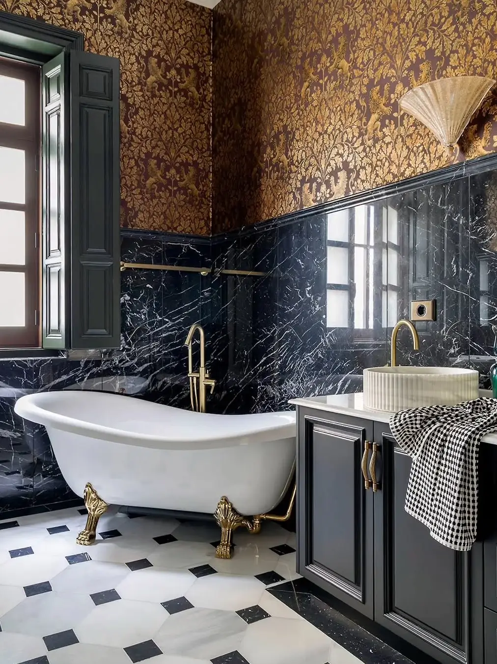 Luxurious bathroom in a restored Murcia mansion featuring a freestanding clawfoot tub, black marble walls, ornate patterned wallpaper, brass fixtures, and geometric marble flooring.