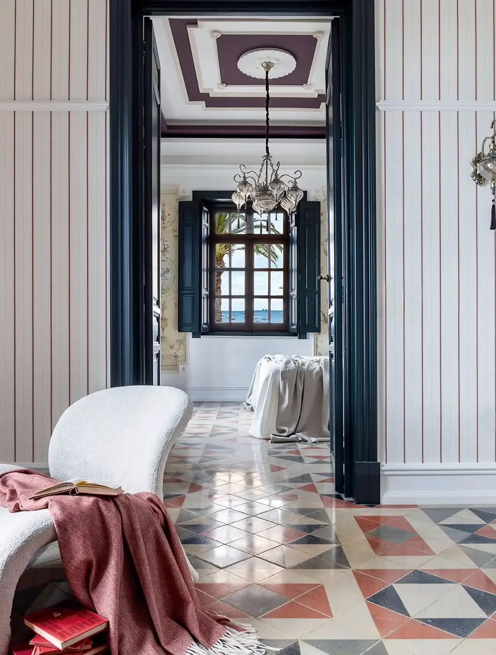 Hallway in a restored Murcia mansion with geometric patterned tile flooring, classic wall paneling, a sculptural lounge chair with a throw, and a doorway leading to a bright room with a chandelier