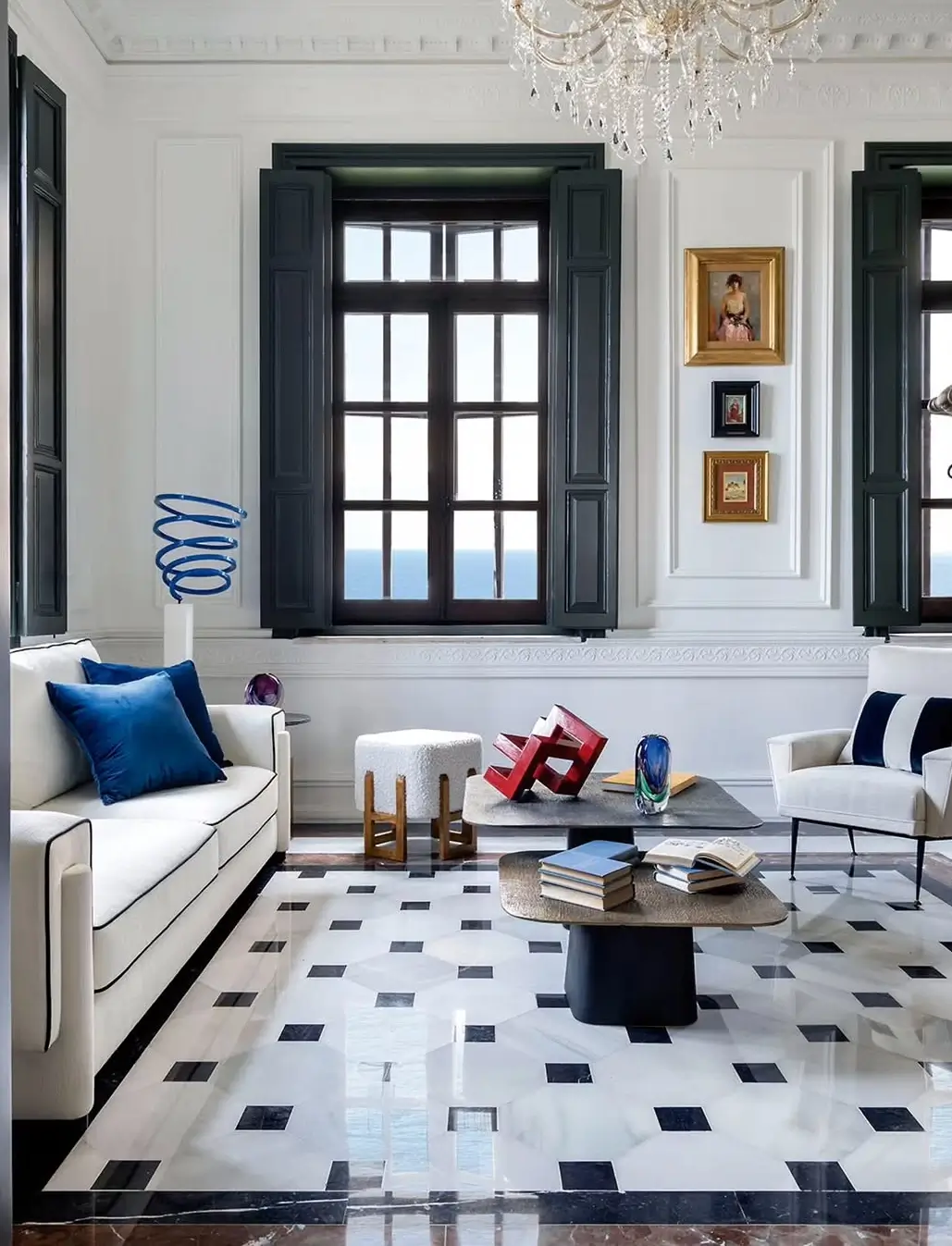 Bright living room in a restored Murcia mansion with black-and-white marble floor, classic wall moldings, modern coffee tables, sculptural decor, blue accent pillows, and large windows with dark shutters.