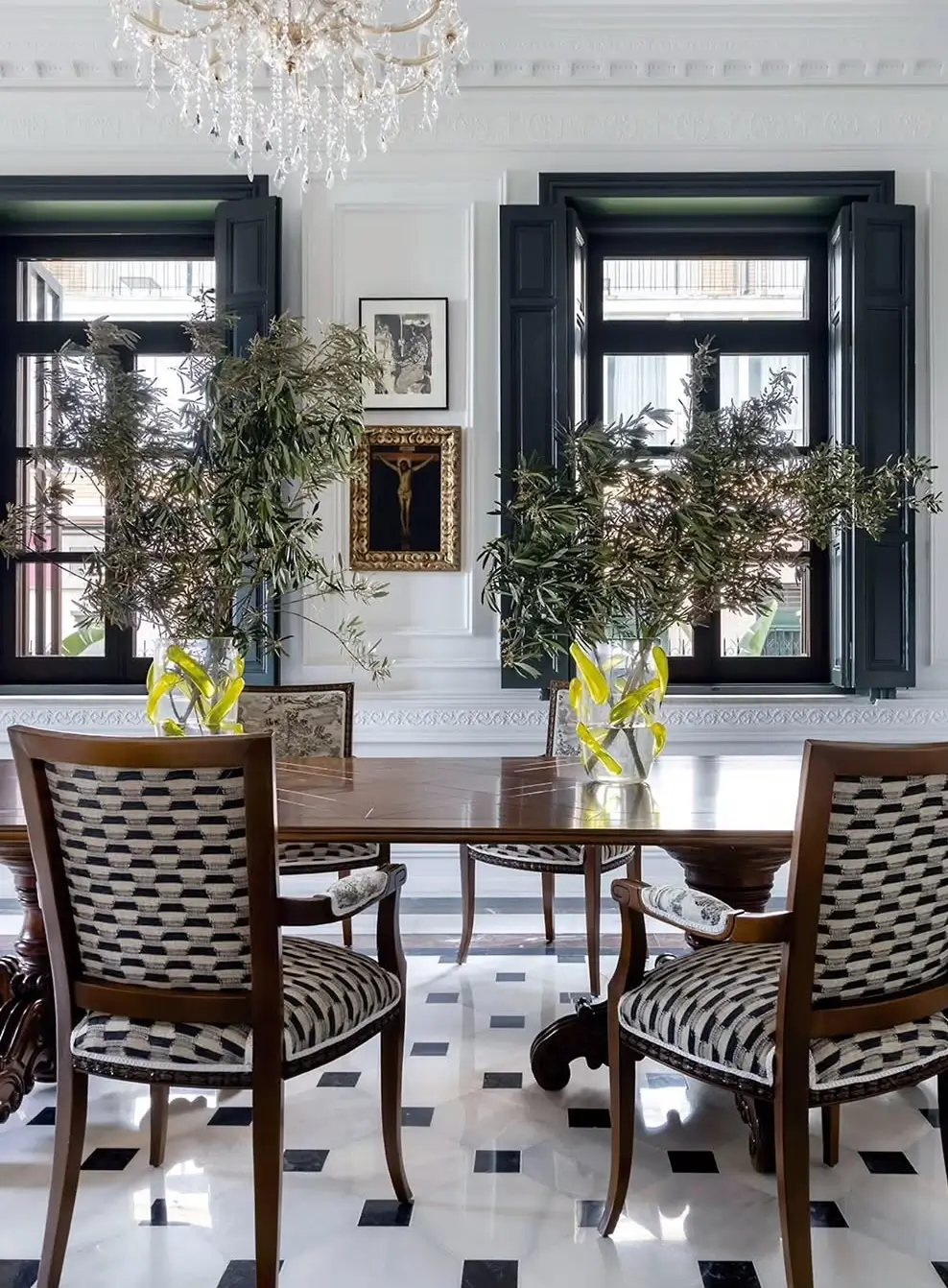 Classic dining room in a restored Murcia mansion with a round wooden table, patterned upholstered chairs, black-and-white marble flooring, tall windows with dark shutters, and fresh greenery arrangements.