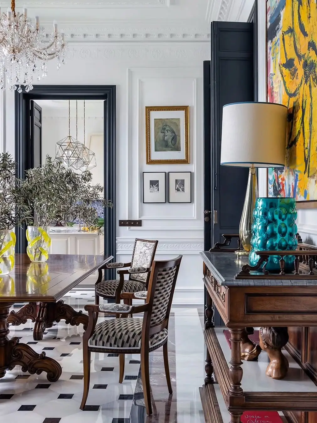 Elegant dining room in a renovated Murcia mansion featuring carved wooden table and chairs, marble geometric flooring, classic wall moldings, layered artwork, and a mix of vintage furniture with colorful modern accents.