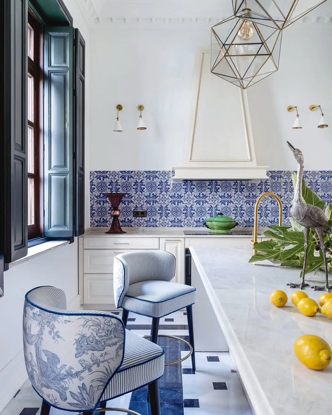 Bright Mediterranean kitchen in a renovated Murcia mansion with blue patterned tile backsplash, marble island, brass fixtures, upholstered bar stools, and large windows with dark shutters.