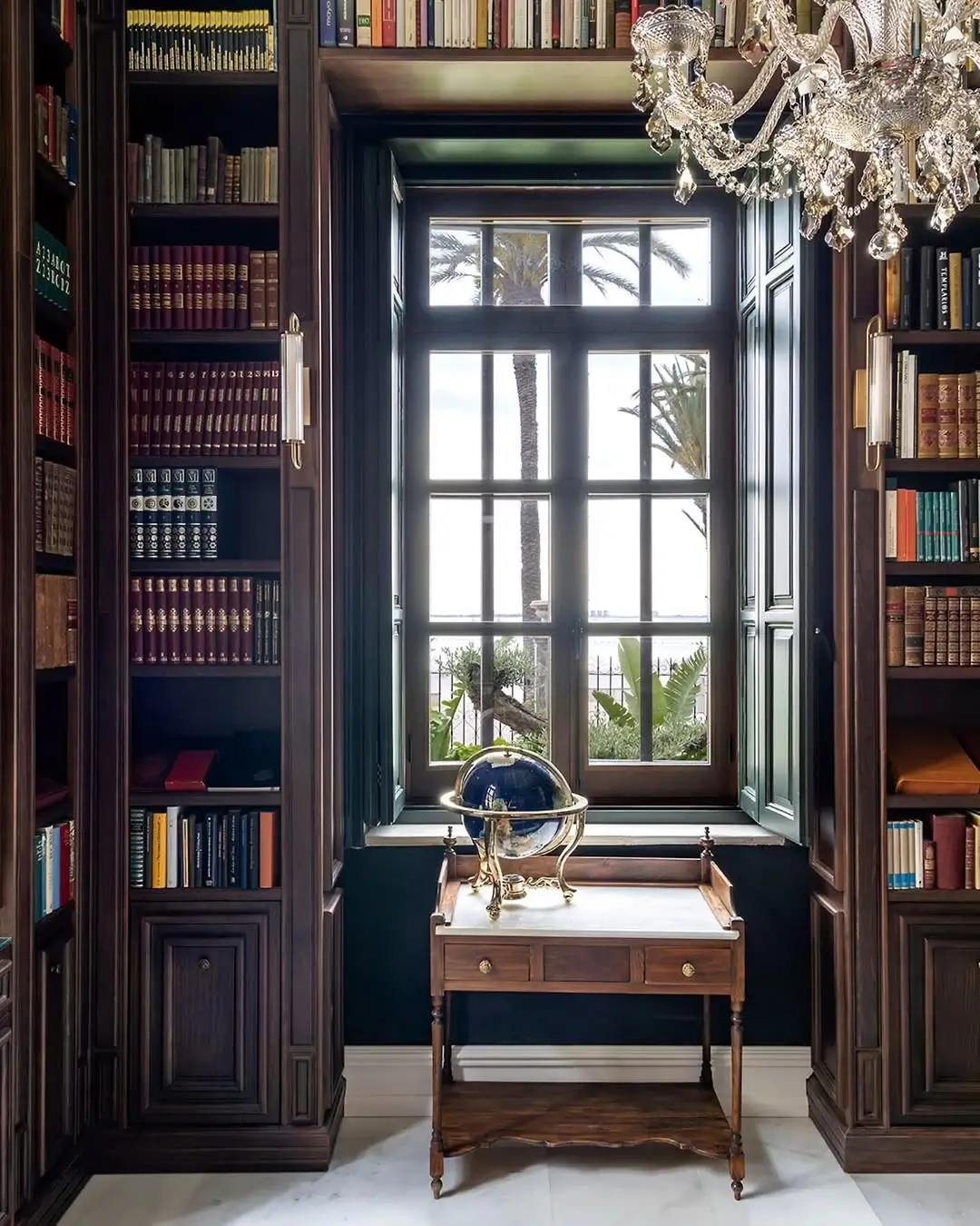 Moody home library in a restored Murcia mansion with floor-to-ceiling wooden bookshelves, a crystal chandelier, a classic desk with a globe, and tall windows framing palm trees outside.