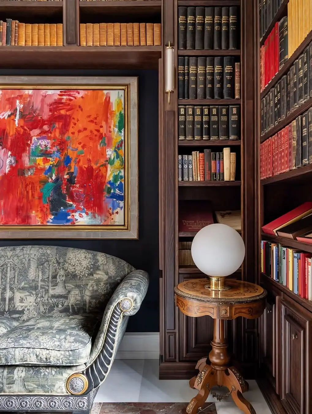 Cozy reading corner in a classic Murcia mansion with built-in wooden bookshelves, an upholstered armchair, a vintage side table with a globe lamp, and a bold abstract painting adding color to the dark walls.