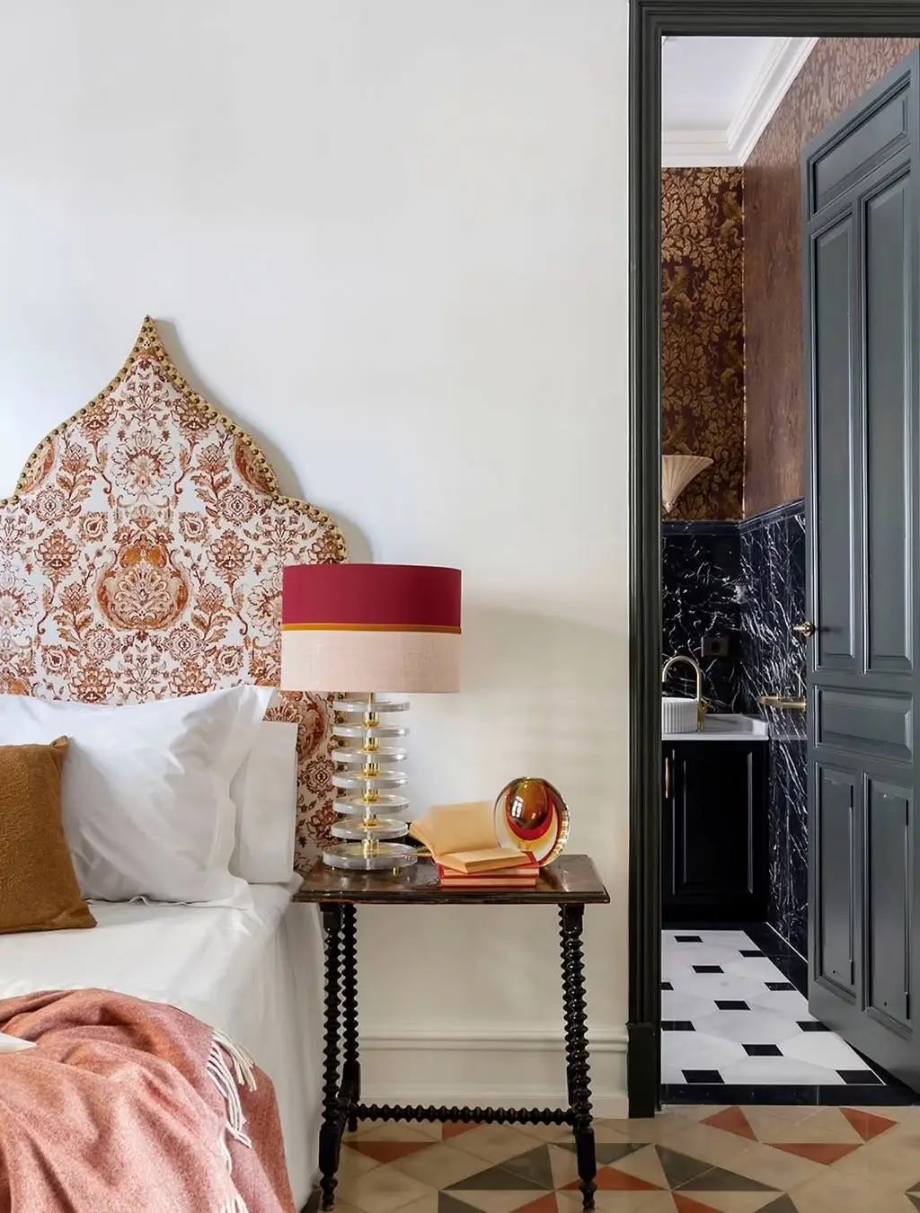 Elegant bedroom in a restored Murcia mansion with a patterned upholstered headboard, vintage bedside table, sculptural lamp, and a view into a dramatic bathroom with dark marble and patterned walls.