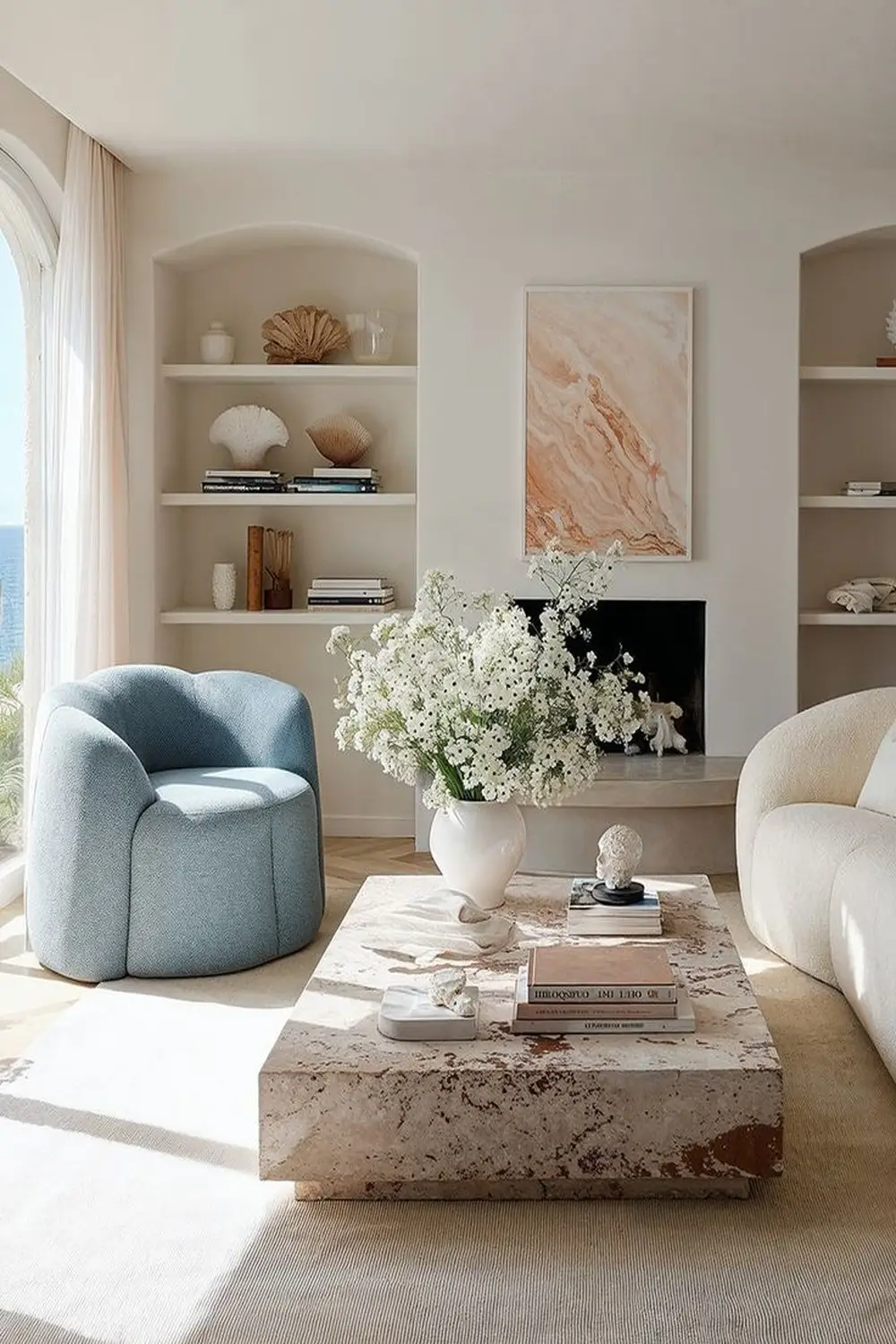 Elegant summer living room with a curved blue armchair, cream sofa, marble coffee table, built-in shelving, and fresh white flowers in a bright coastal-style interior.
Detailed Alt Text: