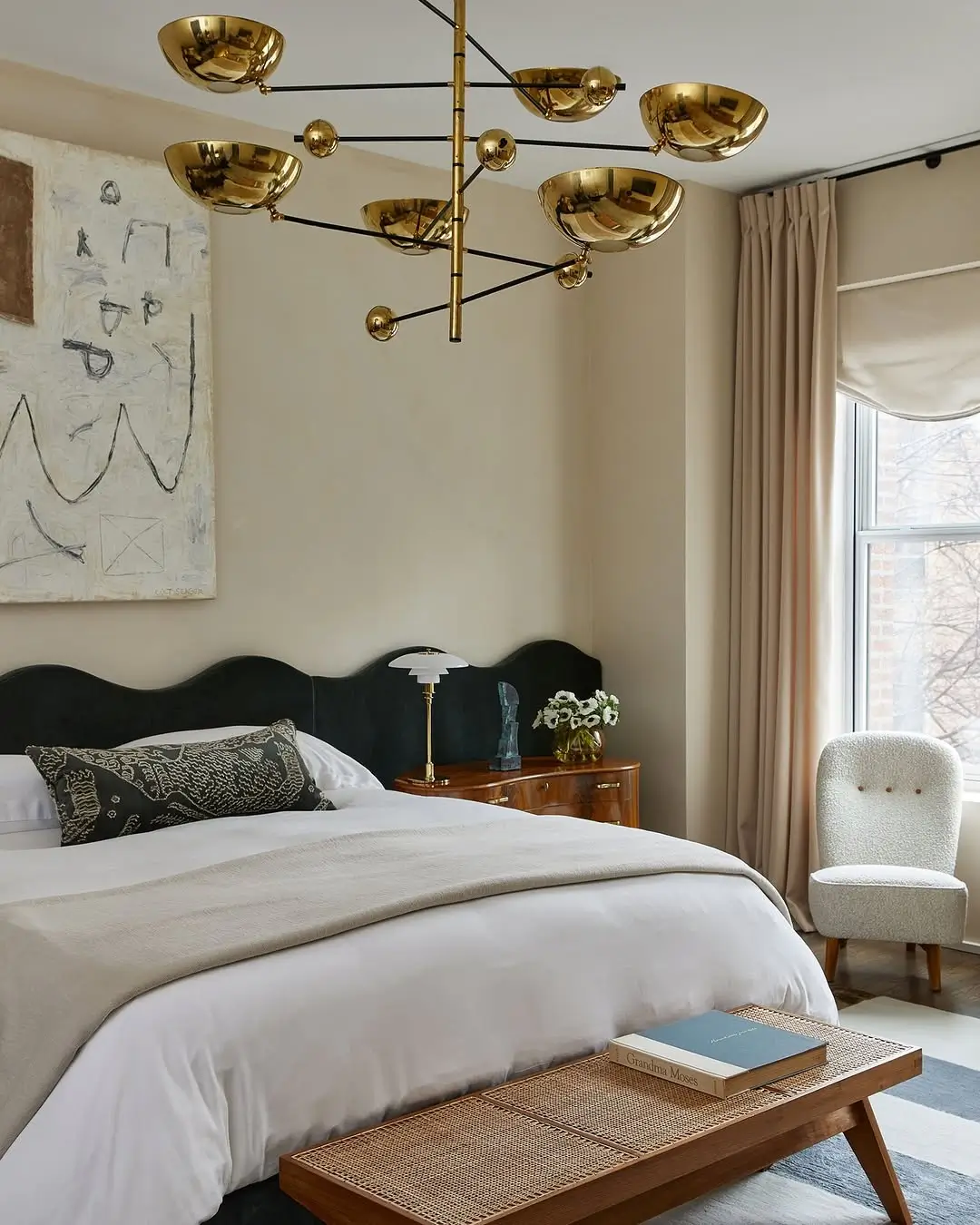 Warm neutral bedroom with creamy beige walls, soft taupe curtains, a sculptural black upholstered headboard, and layered white bedding. Brass lighting, walnut wood furniture, and boucle textures create an elegant, cozy atmosphere with a quiet luxury aesthetic.