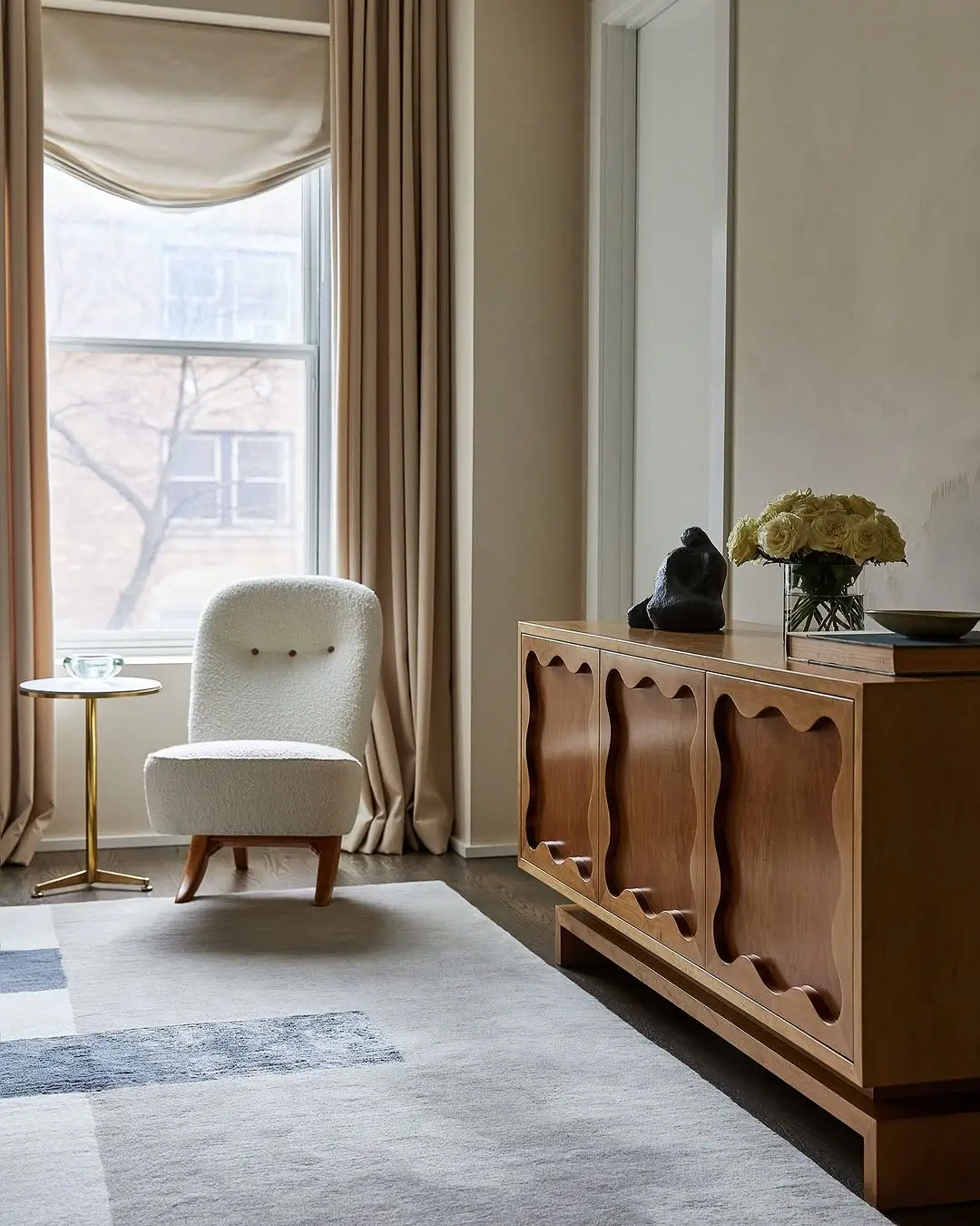 Warm neutral living room with creamy taupe walls, soft beige drapery, a sculptural walnut sideboard, and a boucle accent chair. Natural wood tones, layered textures, and muted earthy colors create a calm, sophisticated atmosphere with a cozy modern feel.