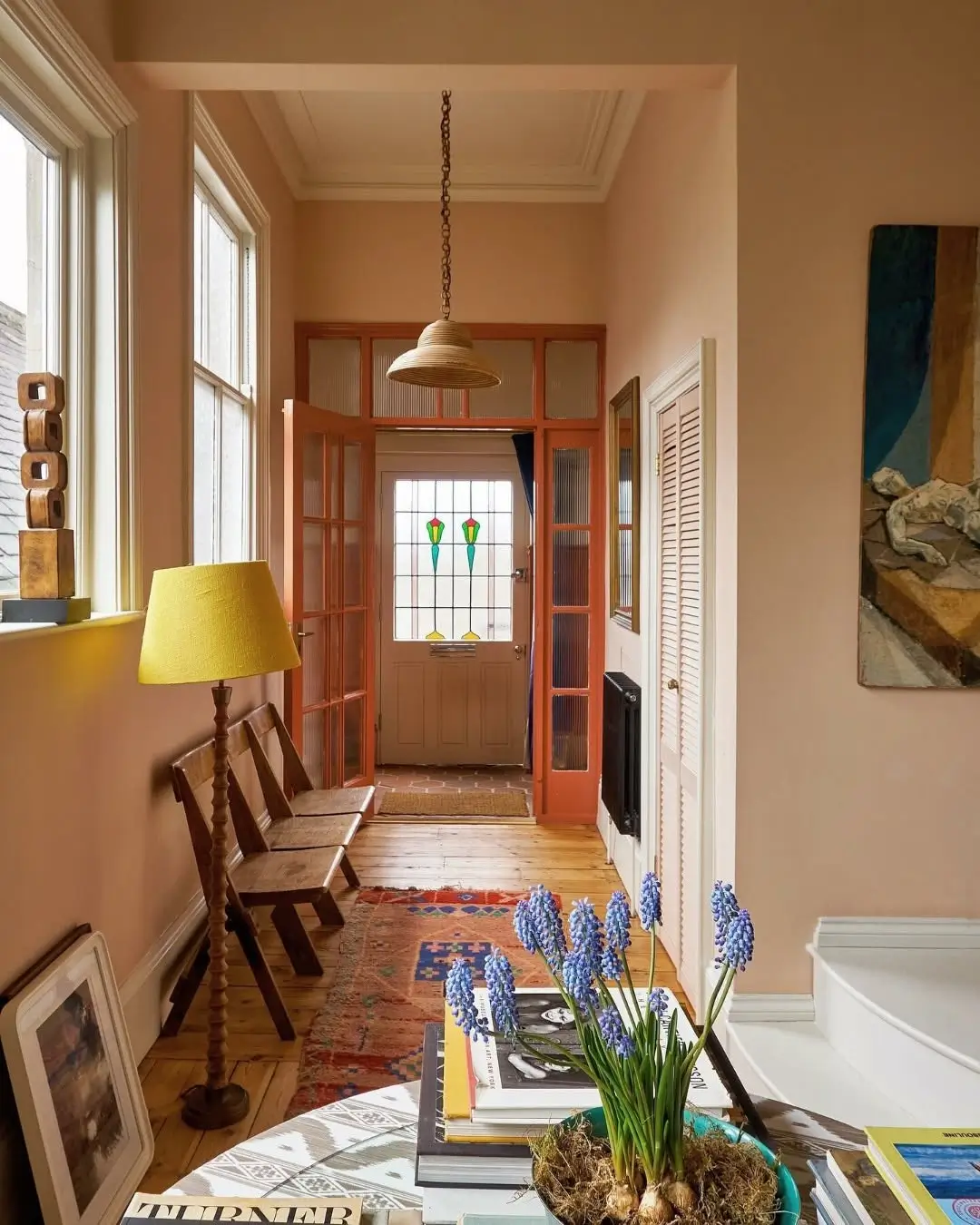 Warm terracotta-toned hallway with peachy clay walls, natural wood floors, vintage wooden chairs, a colorful runner rug, and stained glass front door details. The cozy space features layered earthy tones, soft natural light, sculptural decor, and purple spring flowers styled on a table for a warm, collected atmosphere.