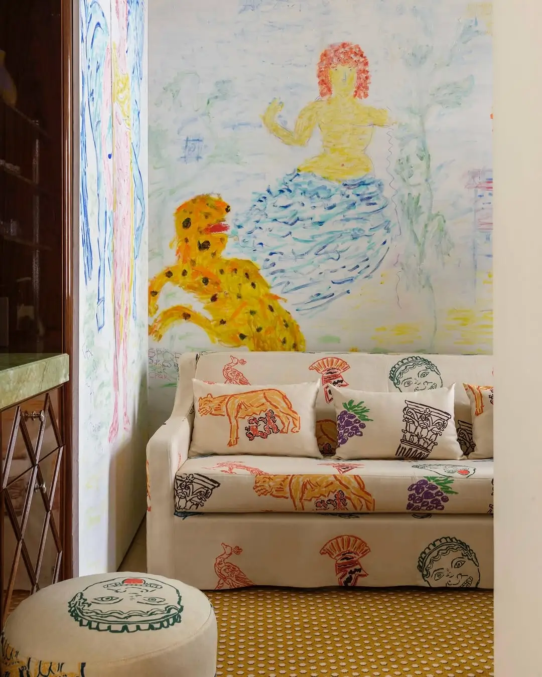 Artist-designed sitting area inside Casa Monti Rome featuring a cream sofa upholstered with playful Roman-inspired illustrations, pastel mural walls with mythological figures, and a whimsical ottoman in Michael McGregor’s signature hand-drawn style.