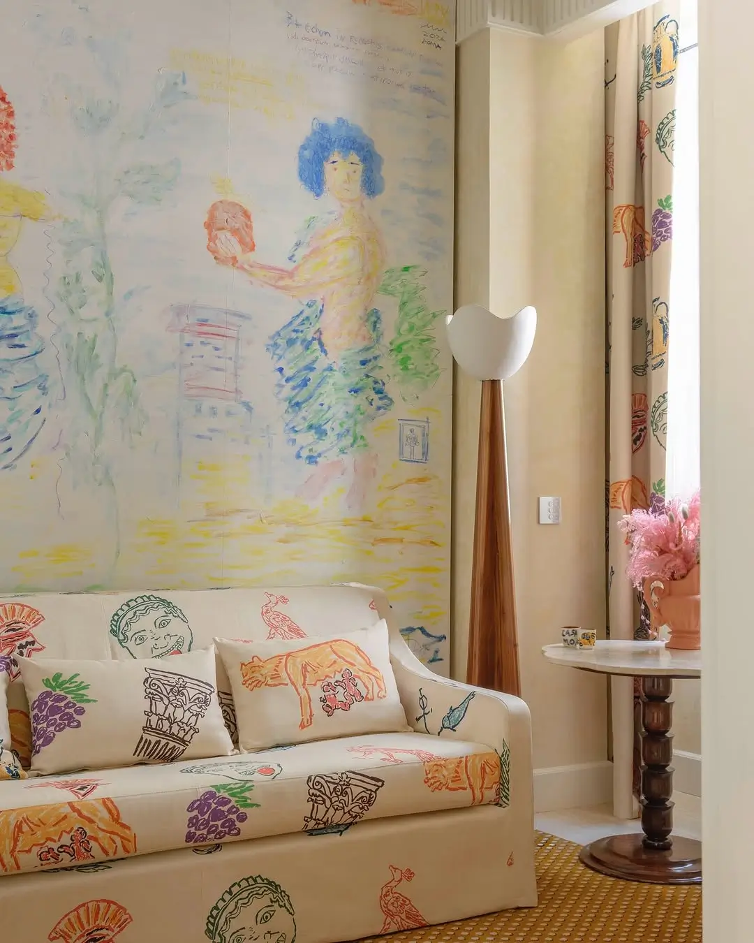 Bright artist-designed lounge area inside Casa Monti Rome featuring a hand-painted pastel mural, illustrated cream sofa with Roman-inspired motifs, sculptural wooden floor lamp, and marble side table with pink flowers.