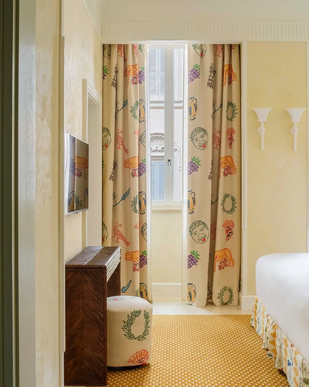 Bedroom inside the Michael McGregor Art Suite at Casa Monti Rome featuring illustrated curtains with Roman-inspired motifs, warm cream walls, patterned carpet, and playful custom textiles in the artist’s signature hand-drawn style.
