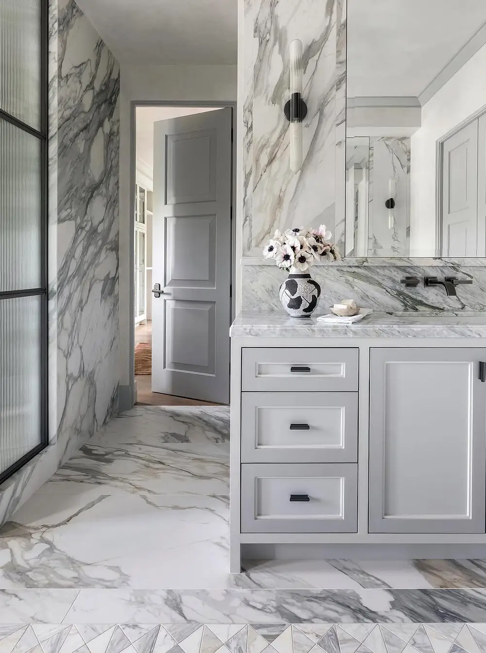 A luxurious modern bathroom clad entirely in dramatic white-and-gray veined marble, creating a sleek spa-like atmosphere. The space features a minimalist gray vanity with recessed panel cabinetry, integrated marble countertops, and matte black wall-mounted faucets. A frameless mirror, sculptural wall sconces, and ribbed glass shower partition add contemporary elegance. Soft gray doors and refined architectural detailing balance the bold marble surfaces, while fresh flowers and curated accessories bring warmth and sophistication to the clean monochromatic palette.