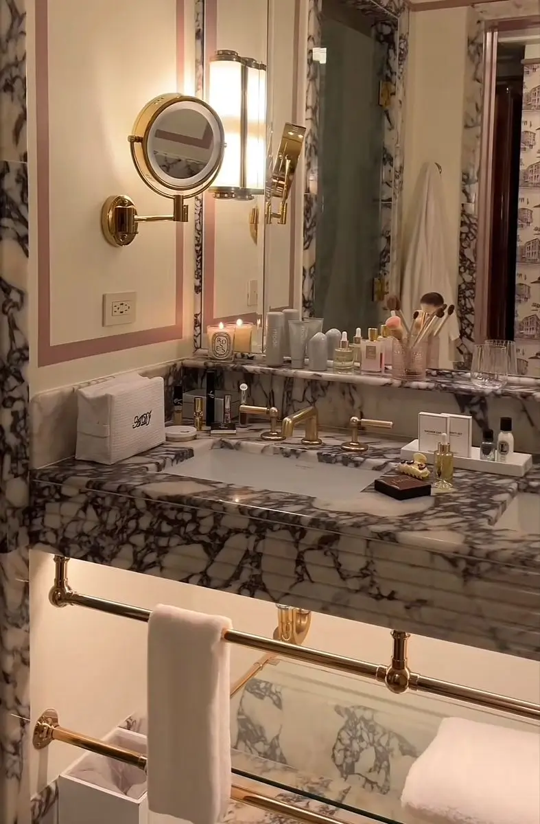 Luxurious hotel bathroom featuring dramatic veined marble walls and countertop, polished brass fixtures, vintage-inspired lighting, and a large mirror lined with beauty products. Soft ambient lighting and elegant finishes create a glamorous boutique hotel atmosphere reminiscent of classic Parisian interiors.