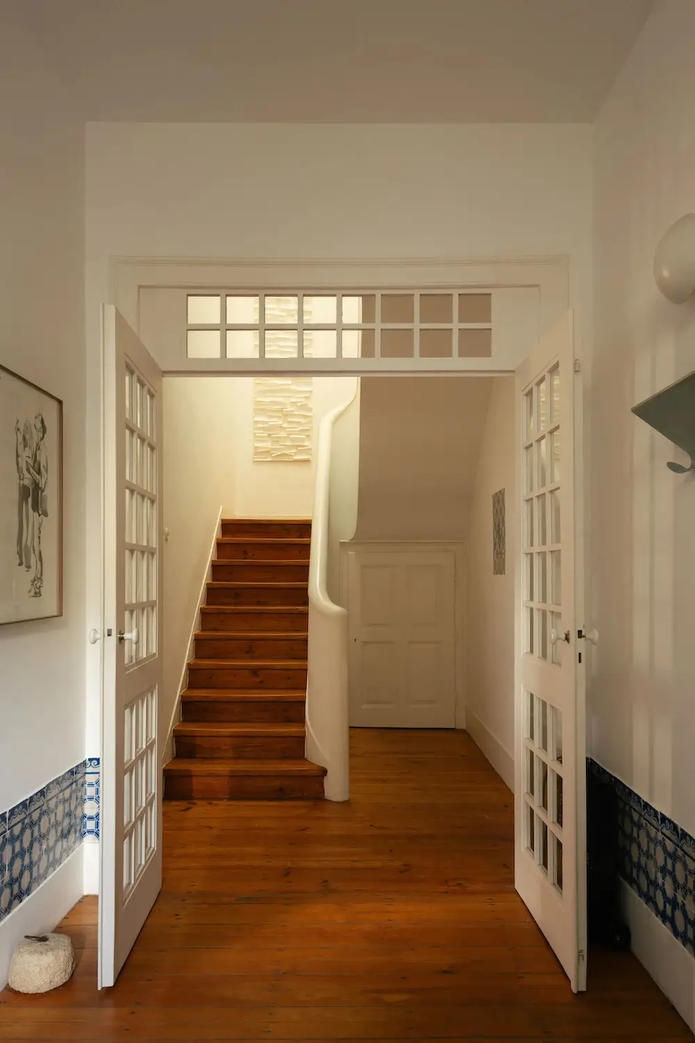 Historic Lisbon townhouse hallway with original pine floors, glass-paneled double doors, blue Portuguese tile details, and a staircase illuminated by natural light.