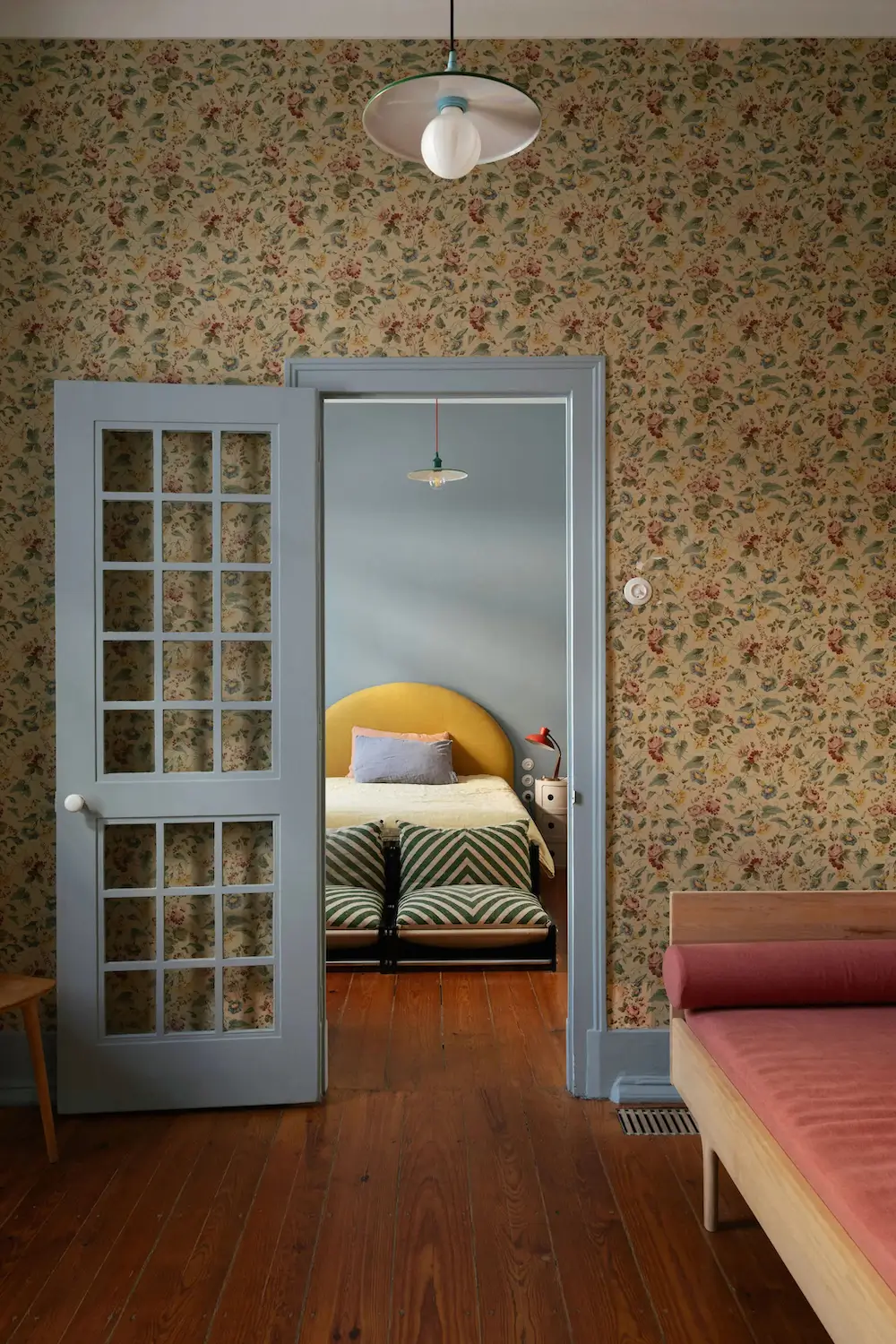 Preserved floral wallpaper frames a view into a blue bedroom with a yellow upholstered headboard, striped floor cushions, and original wood floors in a Lisbon townhouse.