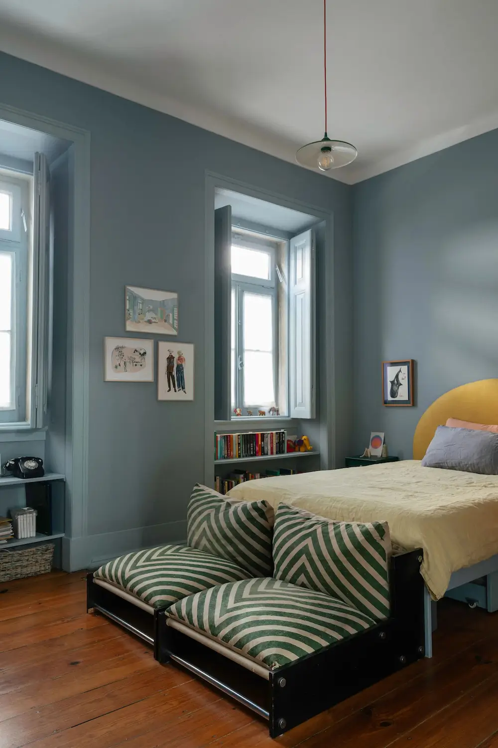 Main bedroom with blue-painted walls, a yellow semicircular headboard, striped floor cushions, built-in bookshelves, and tall traditional windows.