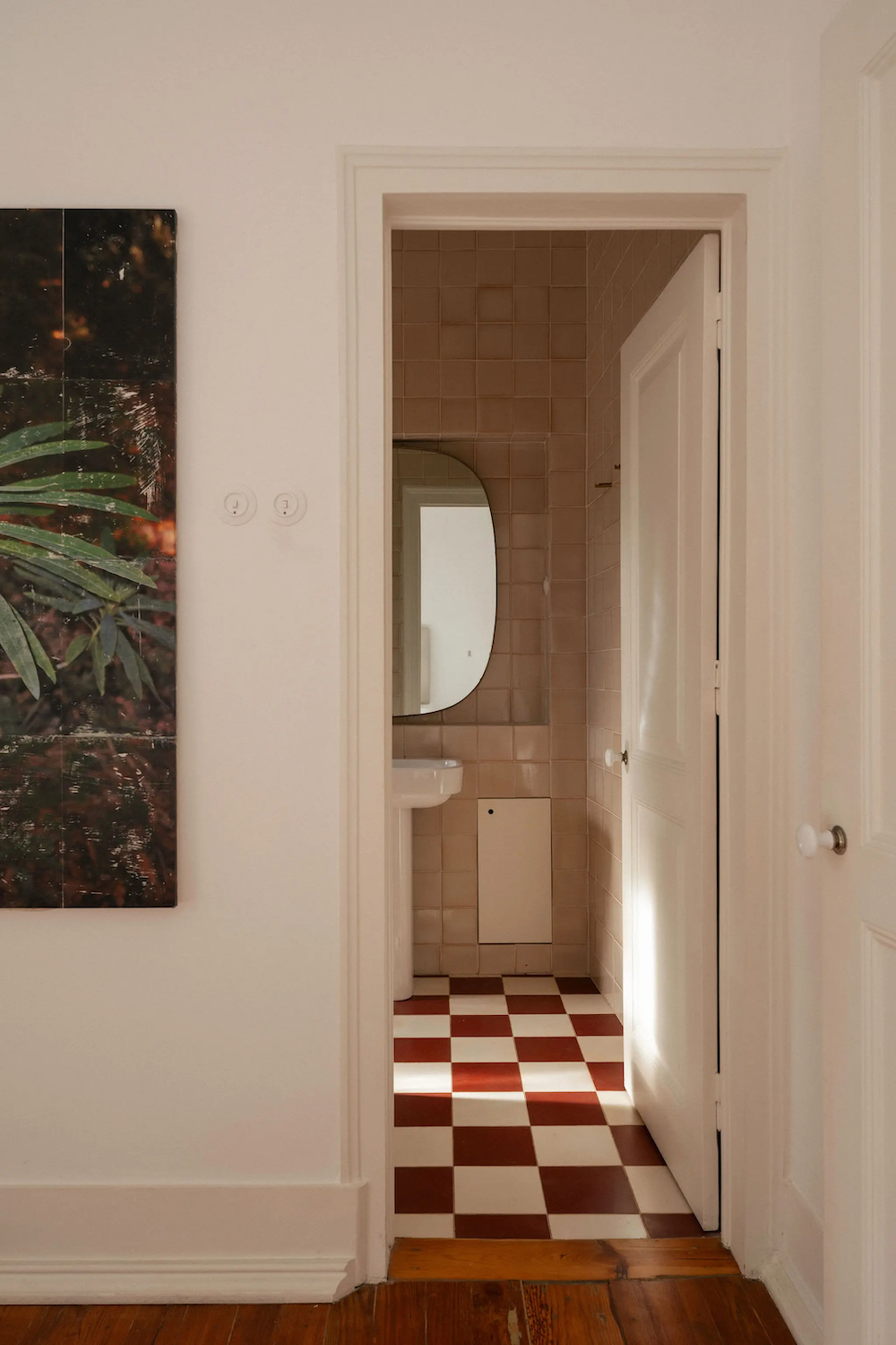 View through a doorway into a compact bathroom featuring blush ceramic wall tiles, a red-and-white checkerboard floor, a pedestal sink, and an arched mirror.