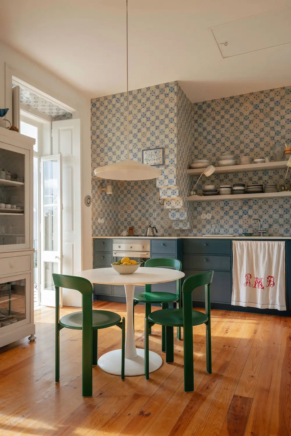 Sunlit Lisbon kitchen featuring original Portuguese azulejo tiles, custom blue cabinets, warm pine floors, and a round white dining table with green chairs.