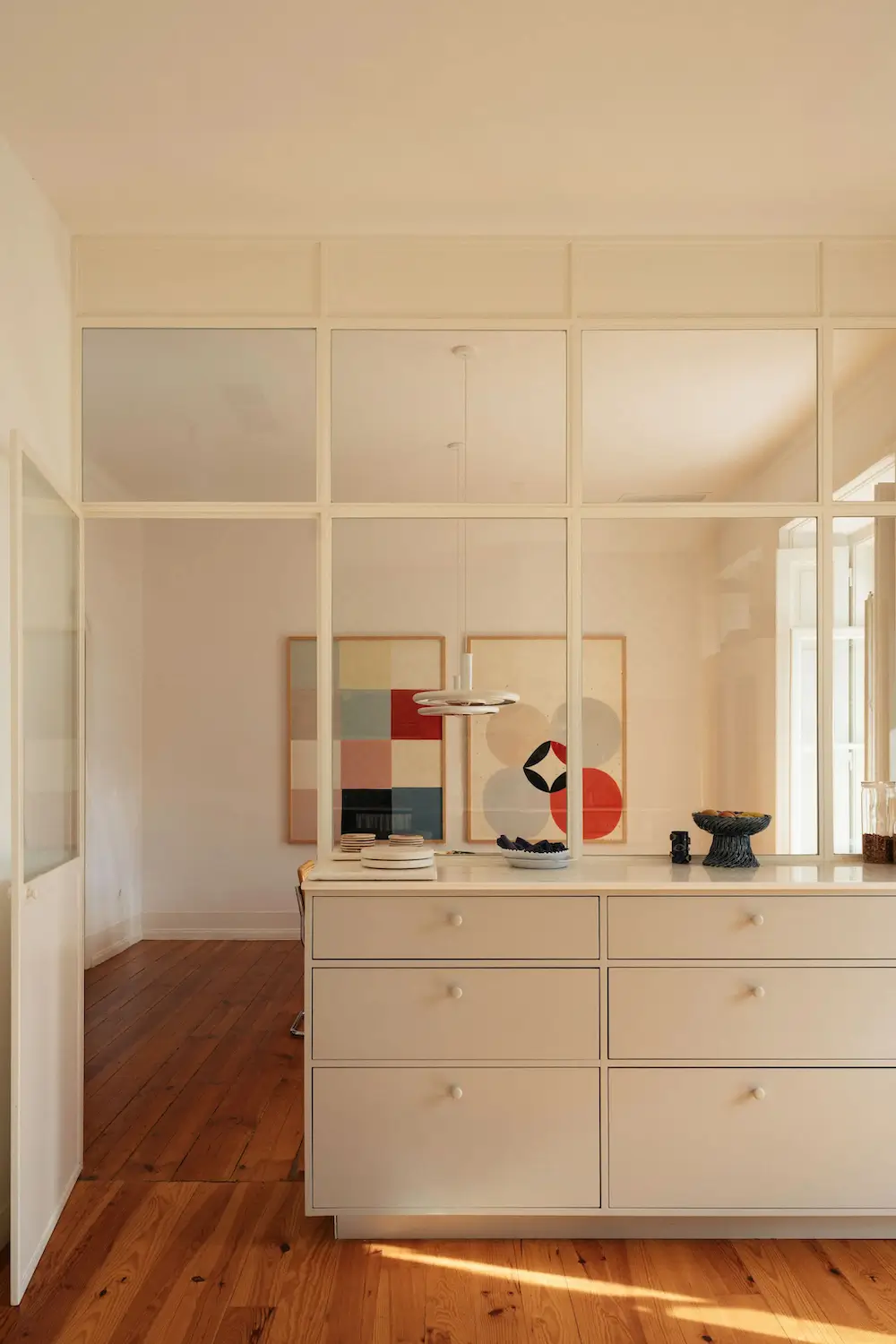 Custom glass partition with integrated storage separates the dining room from the kitchen in a renovated Lisbon townhouse, allowing natural light to flow between spaces.