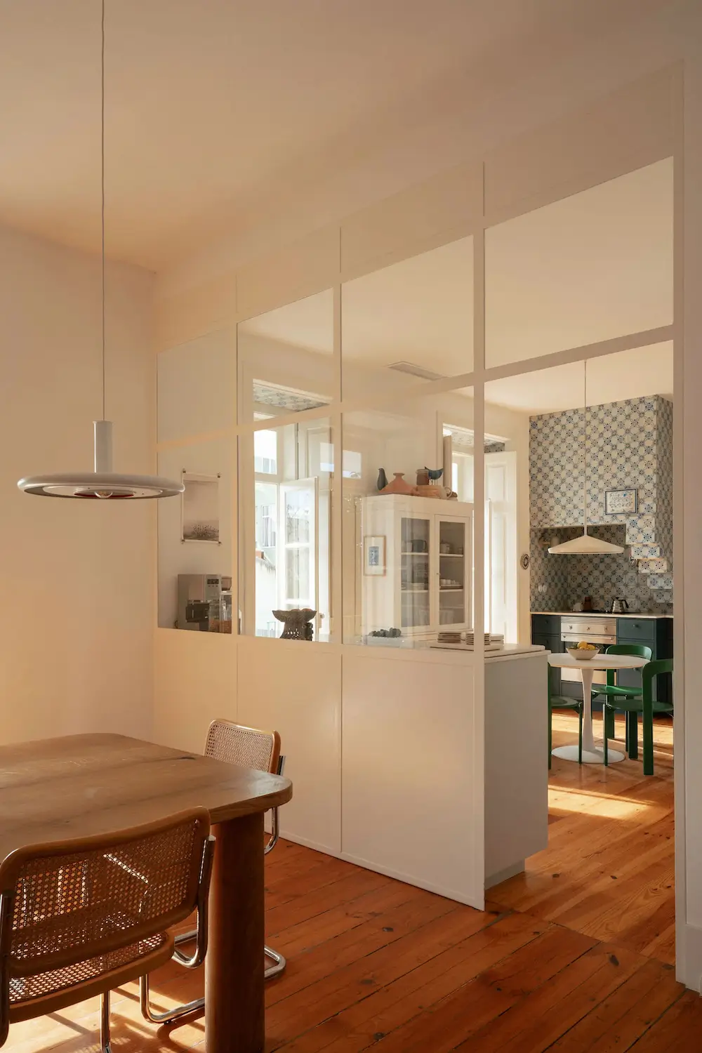 Dining room with a custom wooden table, cane chairs, warm pine floors, and a glass partition overlooking a traditional Portuguese kitchen.