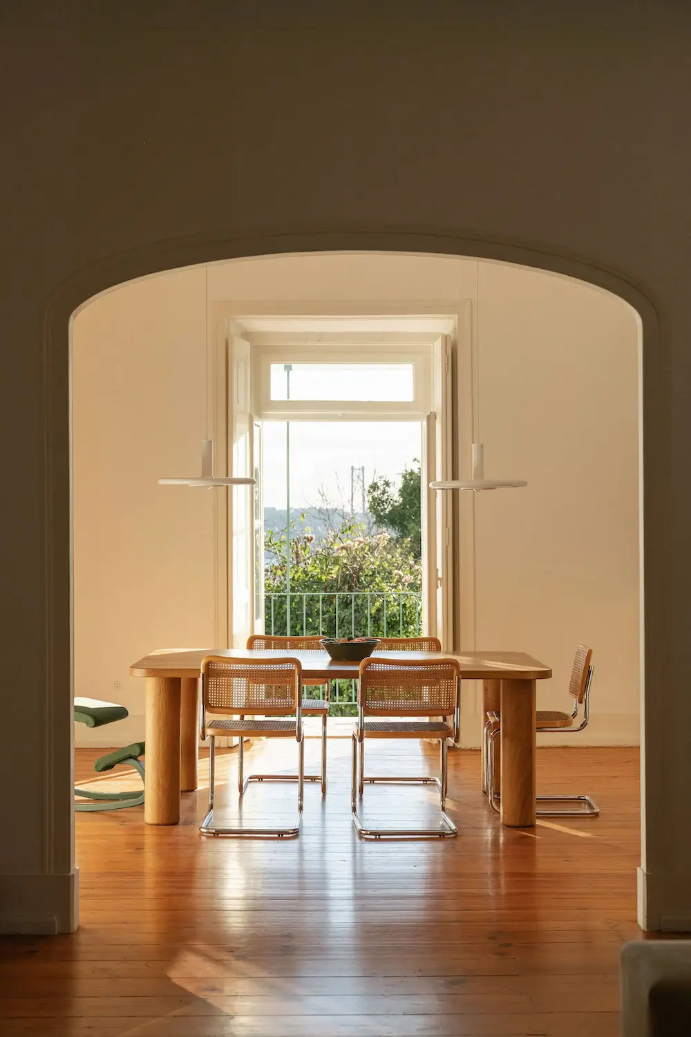 Sunlit dining room framed by a large archway, featuring a custom wooden table, cane chairs, and views toward a balcony in a Lisbon townhouse.
